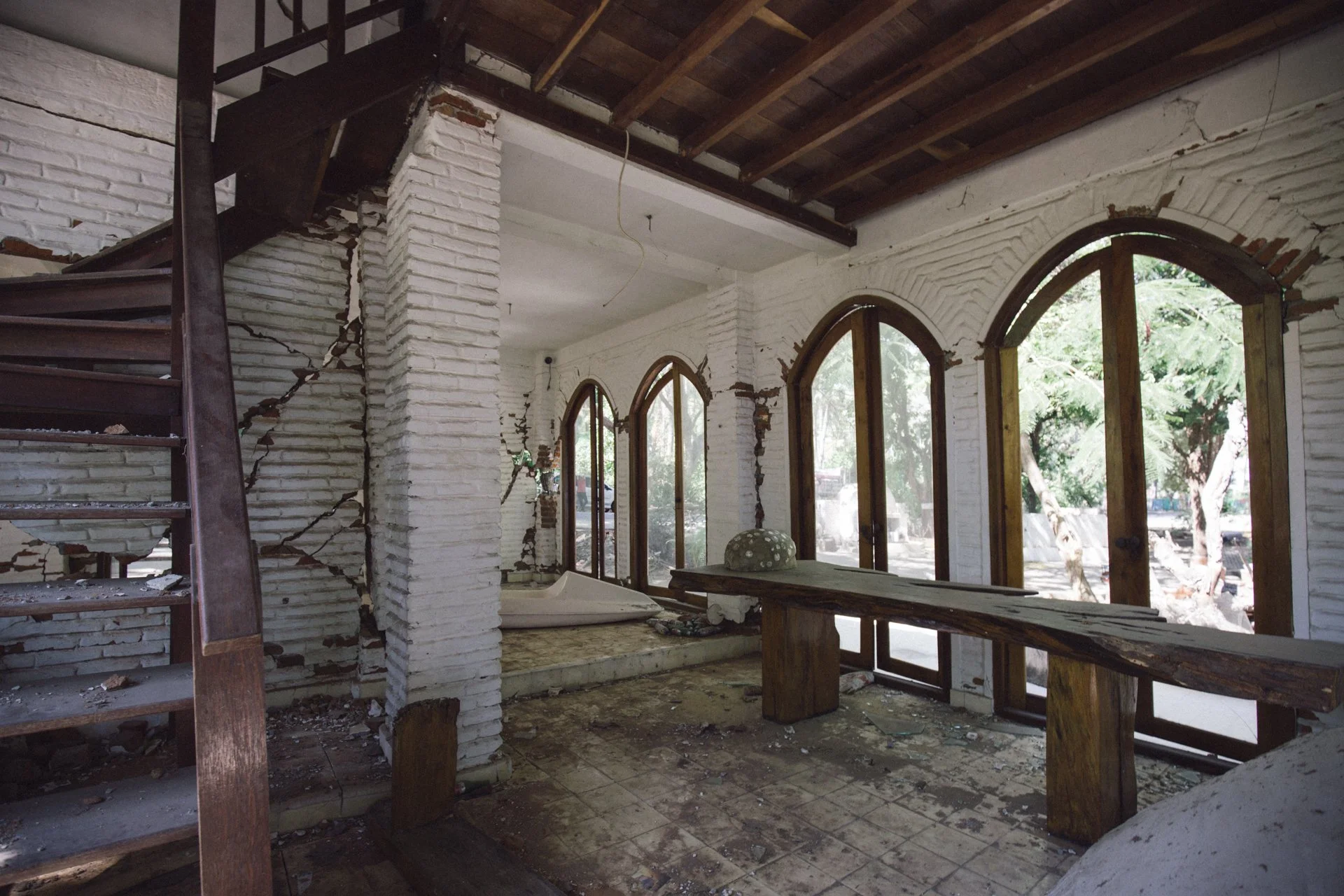 Interior of a building with damaged white brick walls, cracked and broken, exposing inner structure. Large arched windows with wooden frames let in daylight, revealing trees outside. A wooden staircase on the left is partially collapsed. Debris and b