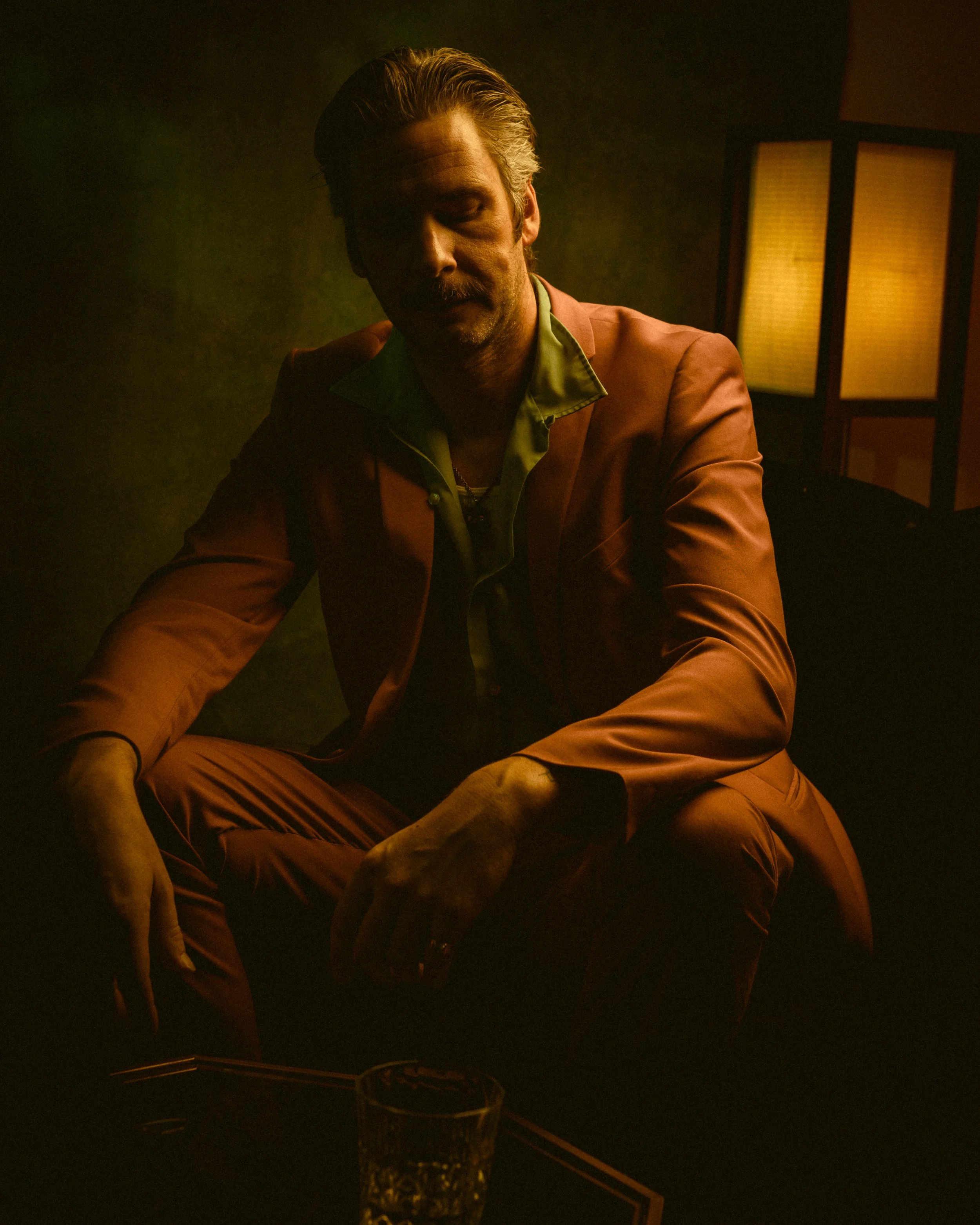 A man with styled hair, a mustache, and beard wears a maroon blazer over a green shirt, sitting in a dimly lit room with a yellow lamp and a glass on the table in front of him.