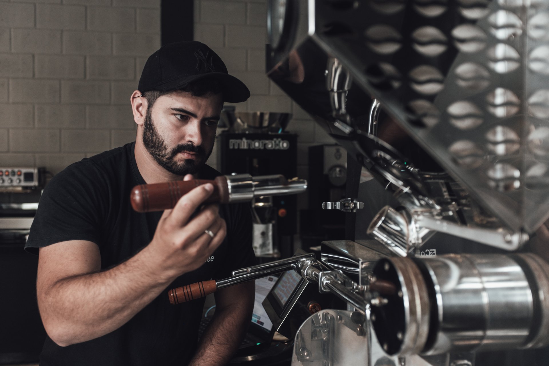A barista with a dark beard and black cap making espresso behind a large commercial espresso machine.