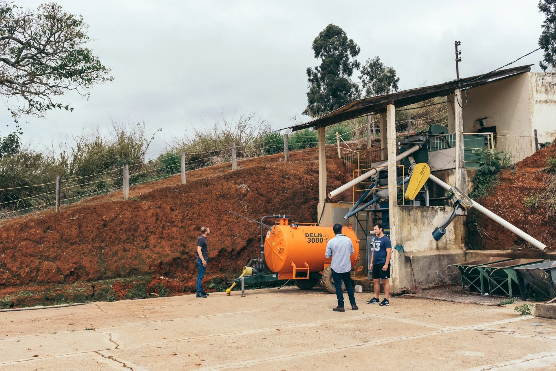 A construction site with four men working near an orange DELN 3000 vacuum truck, with a small building on a hill in the background, trees, and electrical poles.