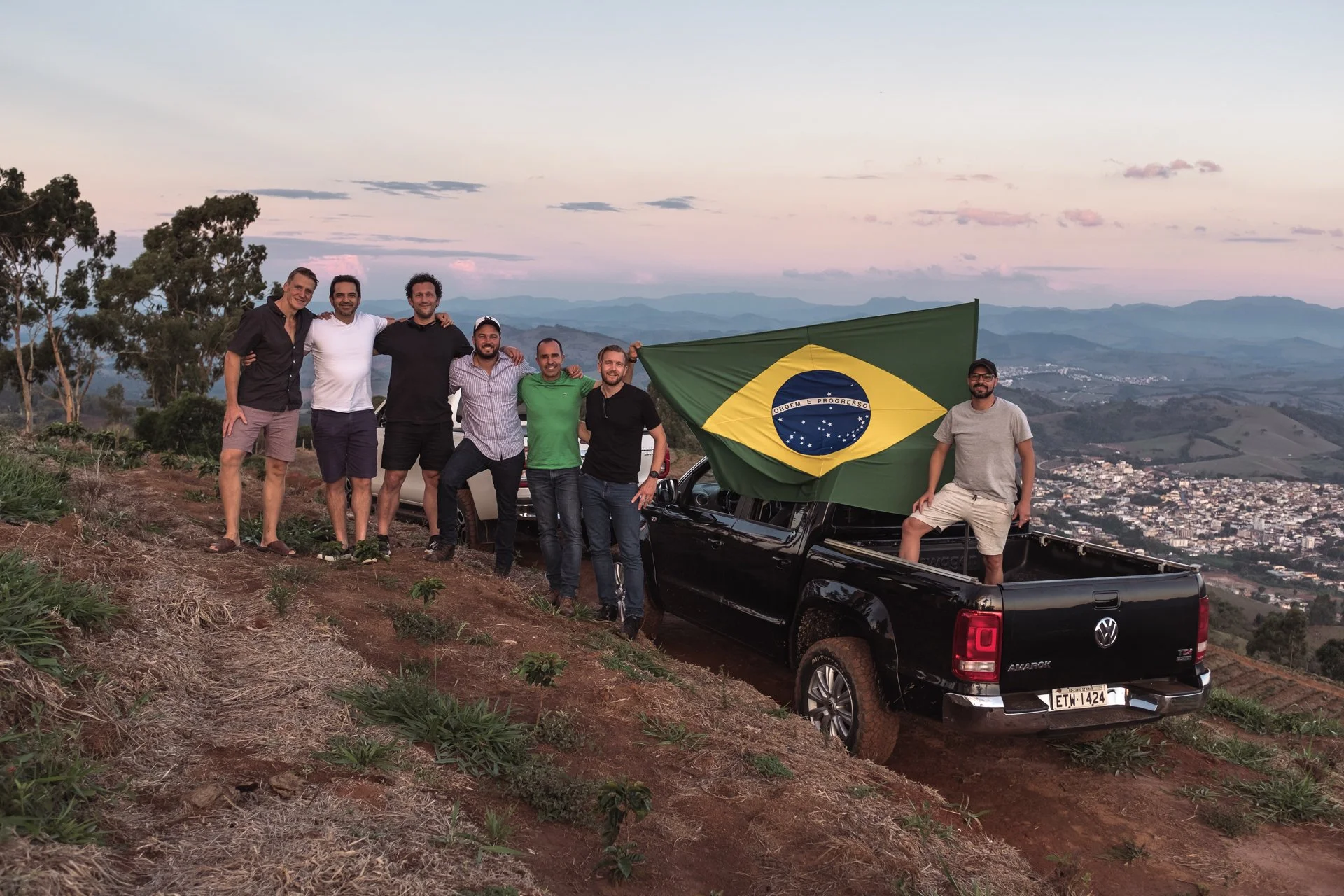Group of seven men posing on a hillside at sunset with a Brazilian flag on a pickup truck, overlooking a valley with houses and mountains.