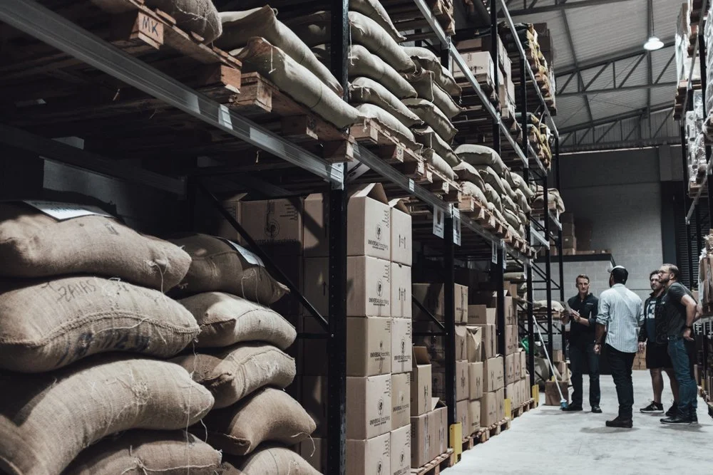 People shopping in a warehouse with shelves filled with bags and boxes.