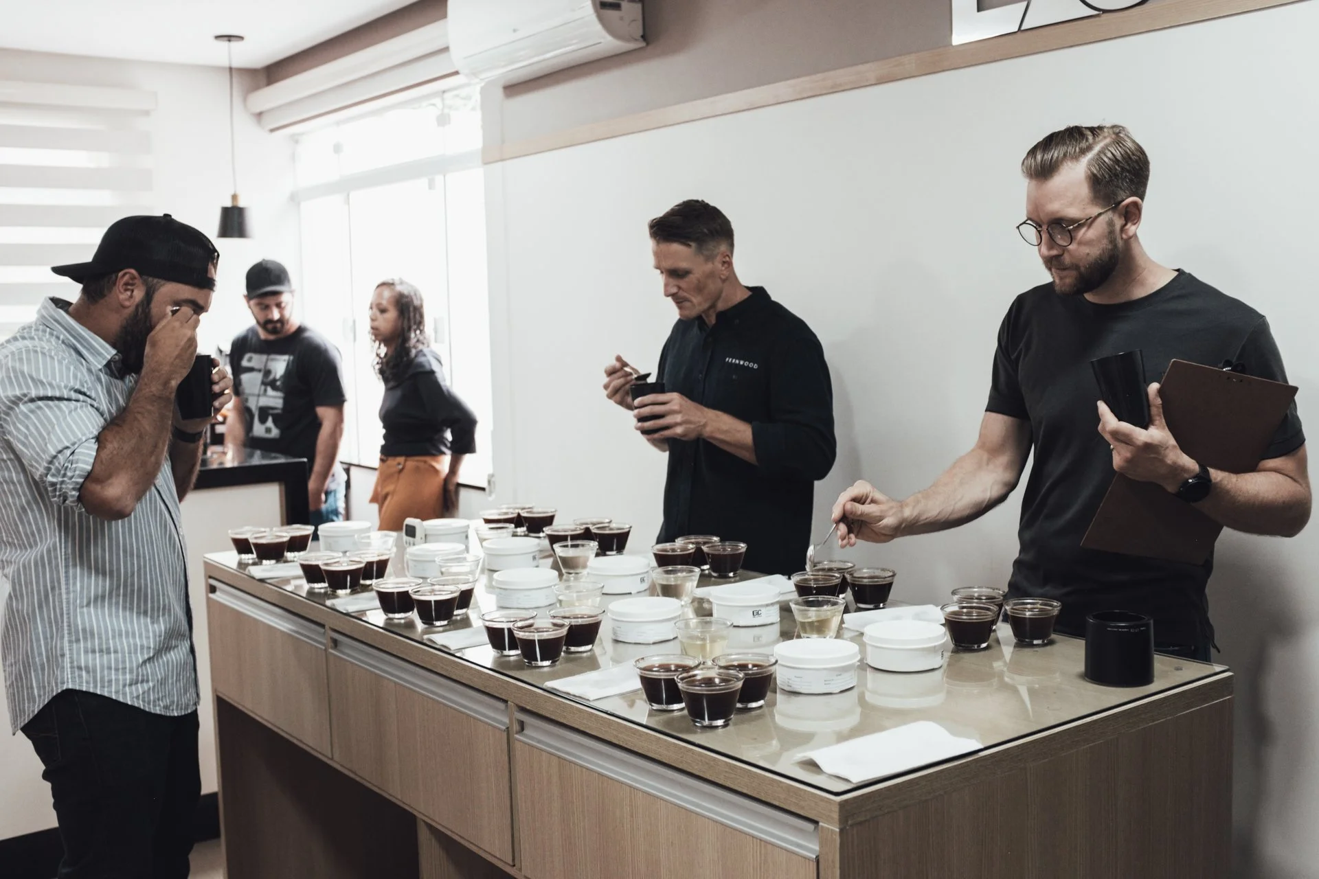 People participating in a coffee tasting event, sampling different types of coffee in small cups arranged on a table in a bright, modern room.