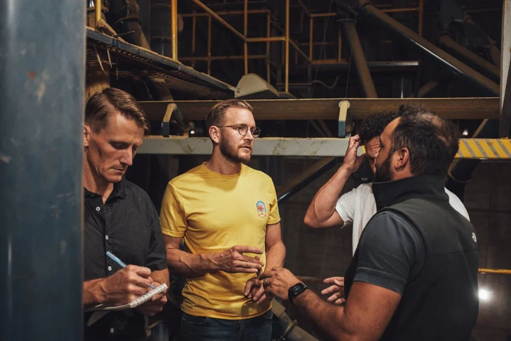 Four men in casual clothing are having a discussion in an industrial or construction setting with metal scaffolding and pipes in the background.
