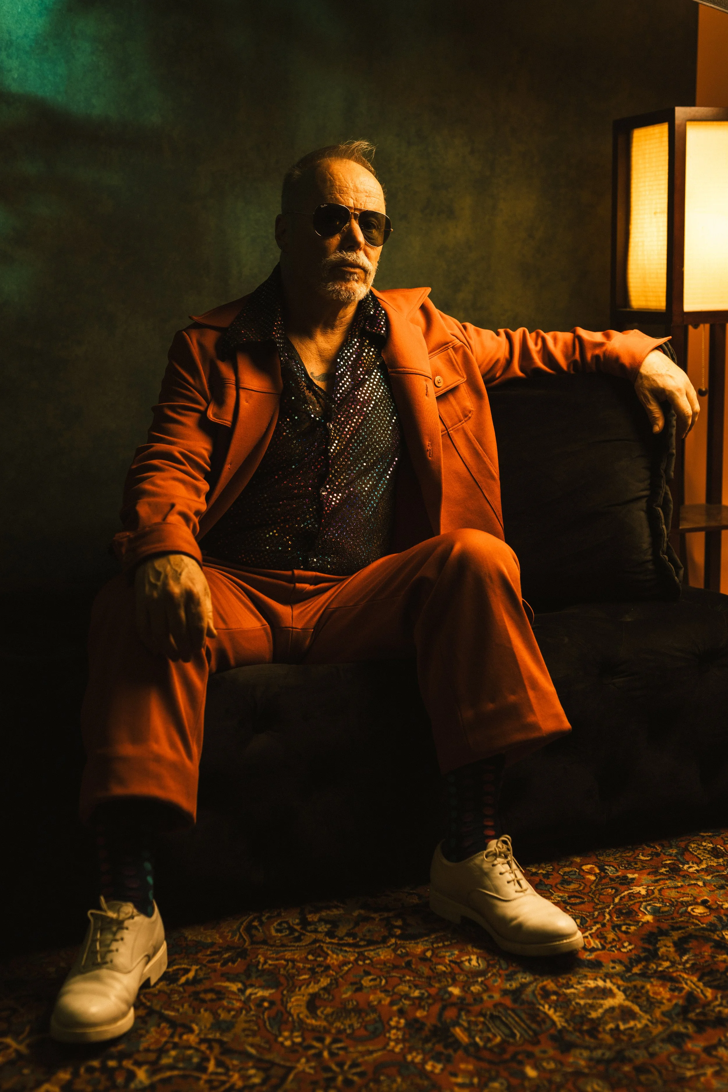 A man with sunglasses, a beard, and a mustache sitting on a black sofa, wearing an orange suit, a sparkly shirt, with his arm resting on the armrest. He is in a dimly lit room with a patterned rug, a floor lamp, and a dark green wall.