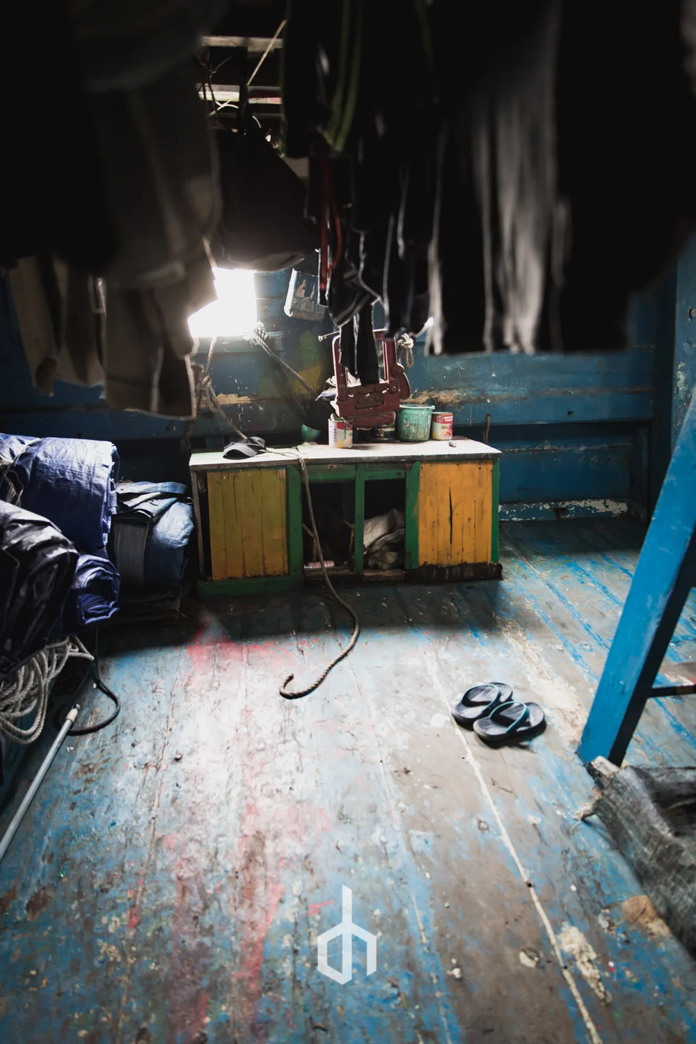 A cluttered room with hanging clothes, a small wooden table with paint cans, a pair of sandals on worn blue and brown painted wooden floor, and a blue ladder to the right.
