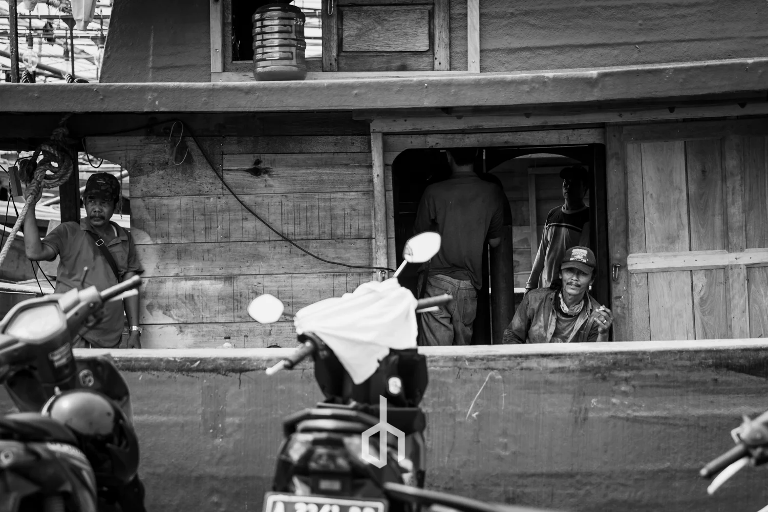 Black and white photo of a rustic wooden building with several men, some inside and some outside. Motorcycles are parked in front, partially blocking the view. The man on the right is sitting and holding a cigarette, while one man on the outside is s