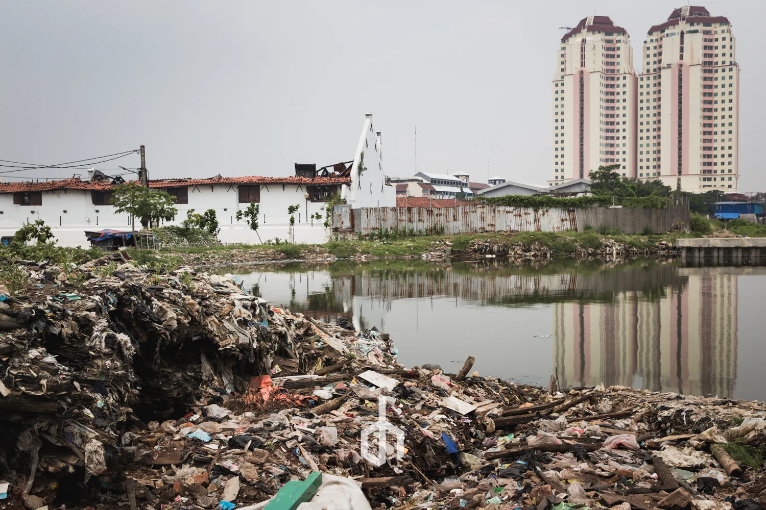 View of a polluted area with trash and debris near a river, with urban buildings and high-rise apartments in the background.