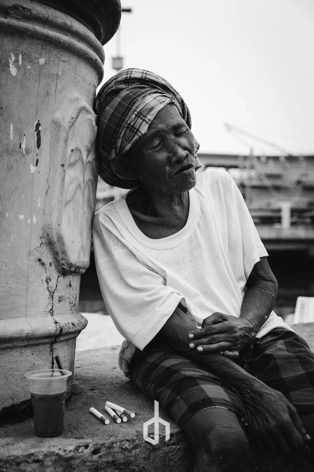 An elderly woman with a headscarf sitting against a large concrete structure, resting her head and eyes closed, near a dock with boats in the background. There are cigarettes and a small cup on the ground in front of her.