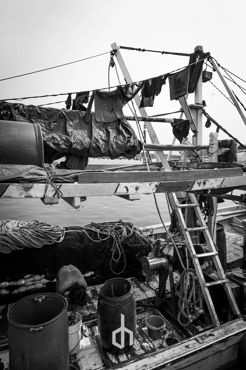 Black and white photo of a boat’s deck with clothes hanging to dry, ropes, barrels, and a ladder, near calm water.