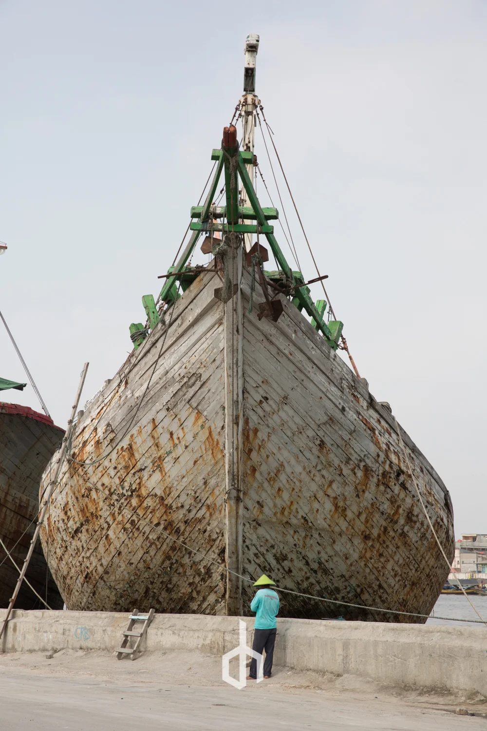 An old, rusty wooden boat is docked on land with a person standing nearby, wearing a conical hat and looking at the boat.