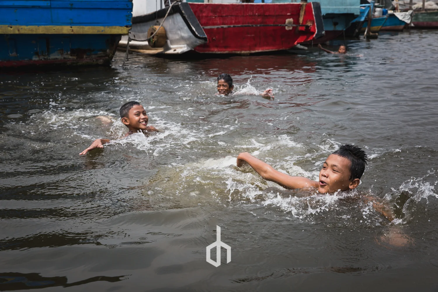 Children swimming and playing in a river, with colorful boats docked along the bank in the background.