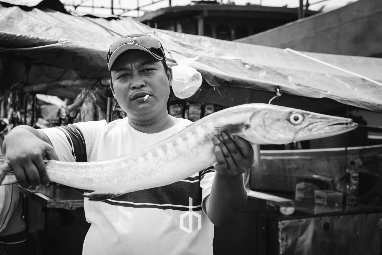A man holding a long, slim fish with a pointed head at a market or fishing dock area. The man has a cigarette in his mouth and is wearing a cap, sunglasses, and a t-shirt. The background shows market stalls or boats.