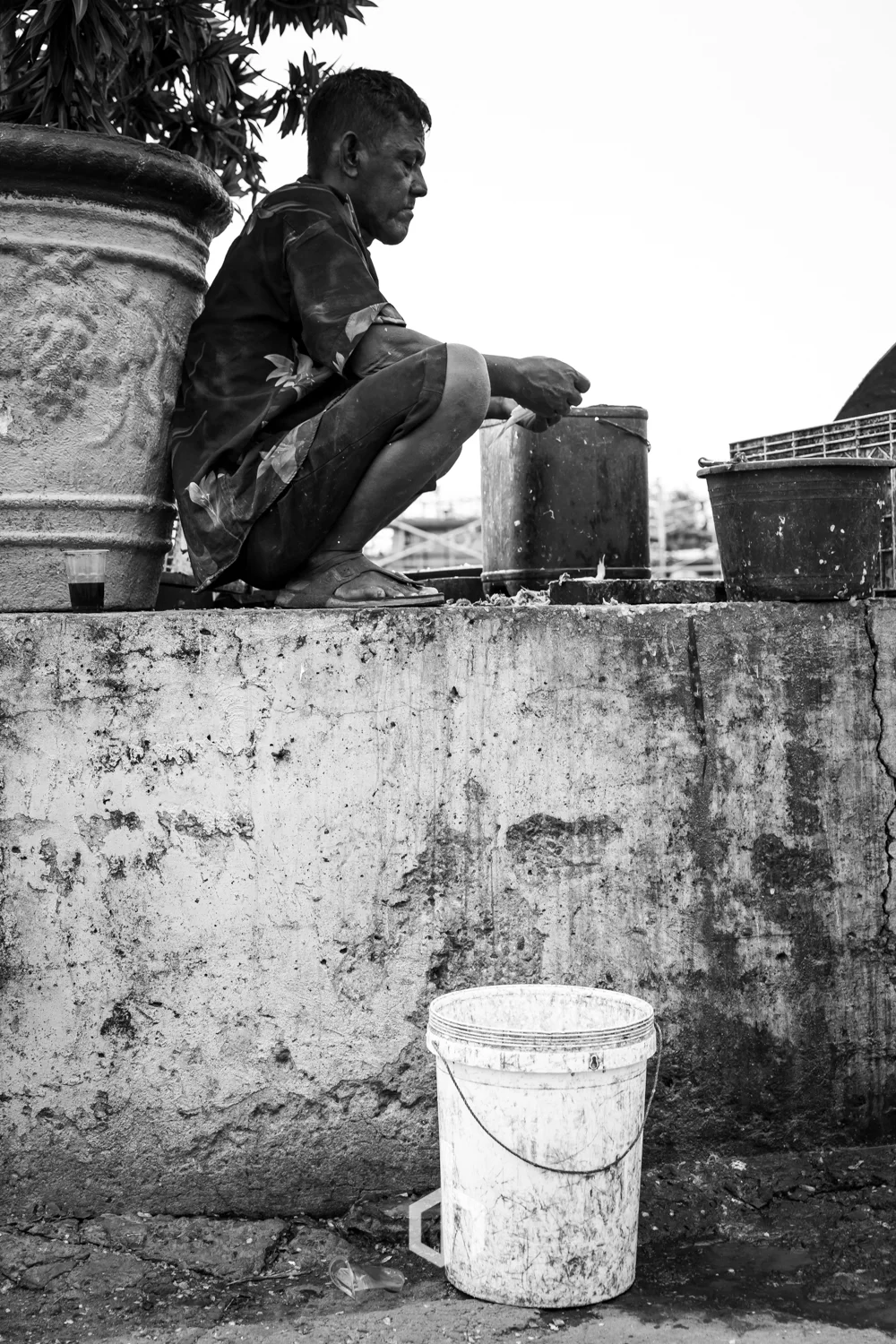A man sitting on a concrete ledge, filling a container with water from a faucet, with a white bucket placed on the ground below. The man appears focused on his task in an outdoor setting.