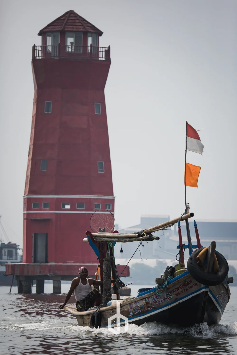 A small boat with a man onboard sailing in front of a tall red lighthouse with a viewing platform at the top, with flags flying on the boat.