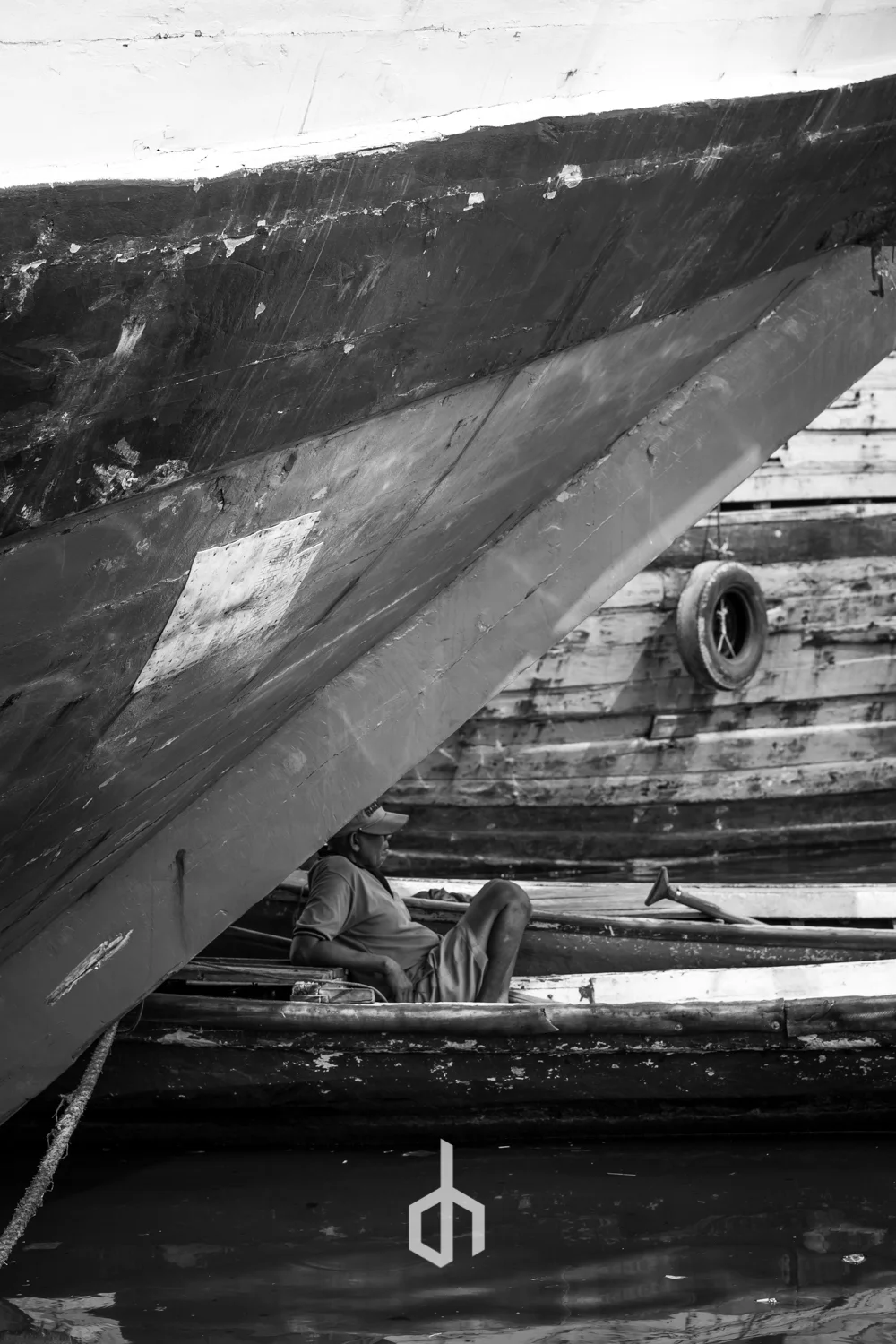 A person sitting in a boat under a large docked vessel, with wooden planks and tires visible in the background, in black and white.