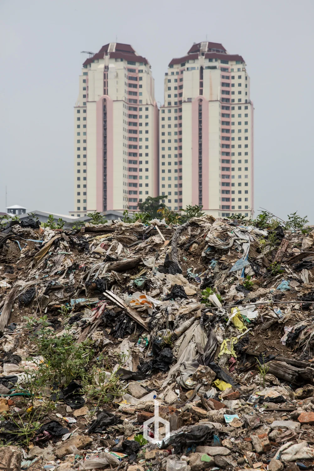 A large pile of garbage and debris in the foreground with two tall, modern apartment buildings in the background under a gray sky.