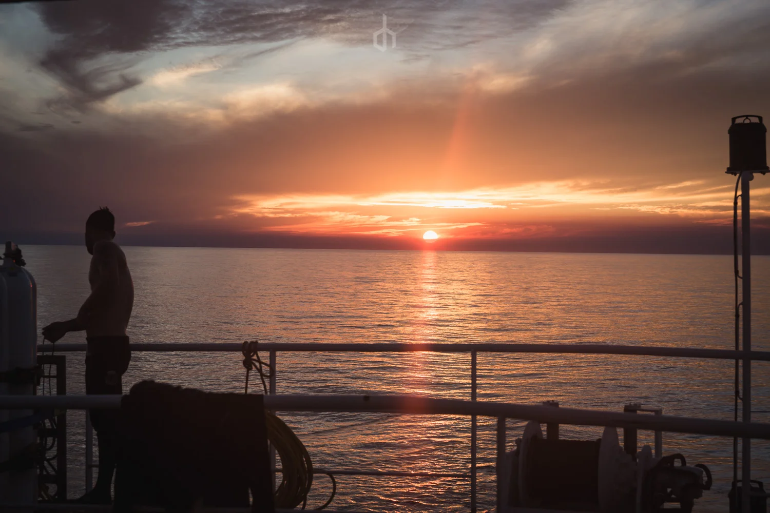 Silhouette of a person on a boat deck during sunset over the ocean, with clouds and colorful sky in the background.