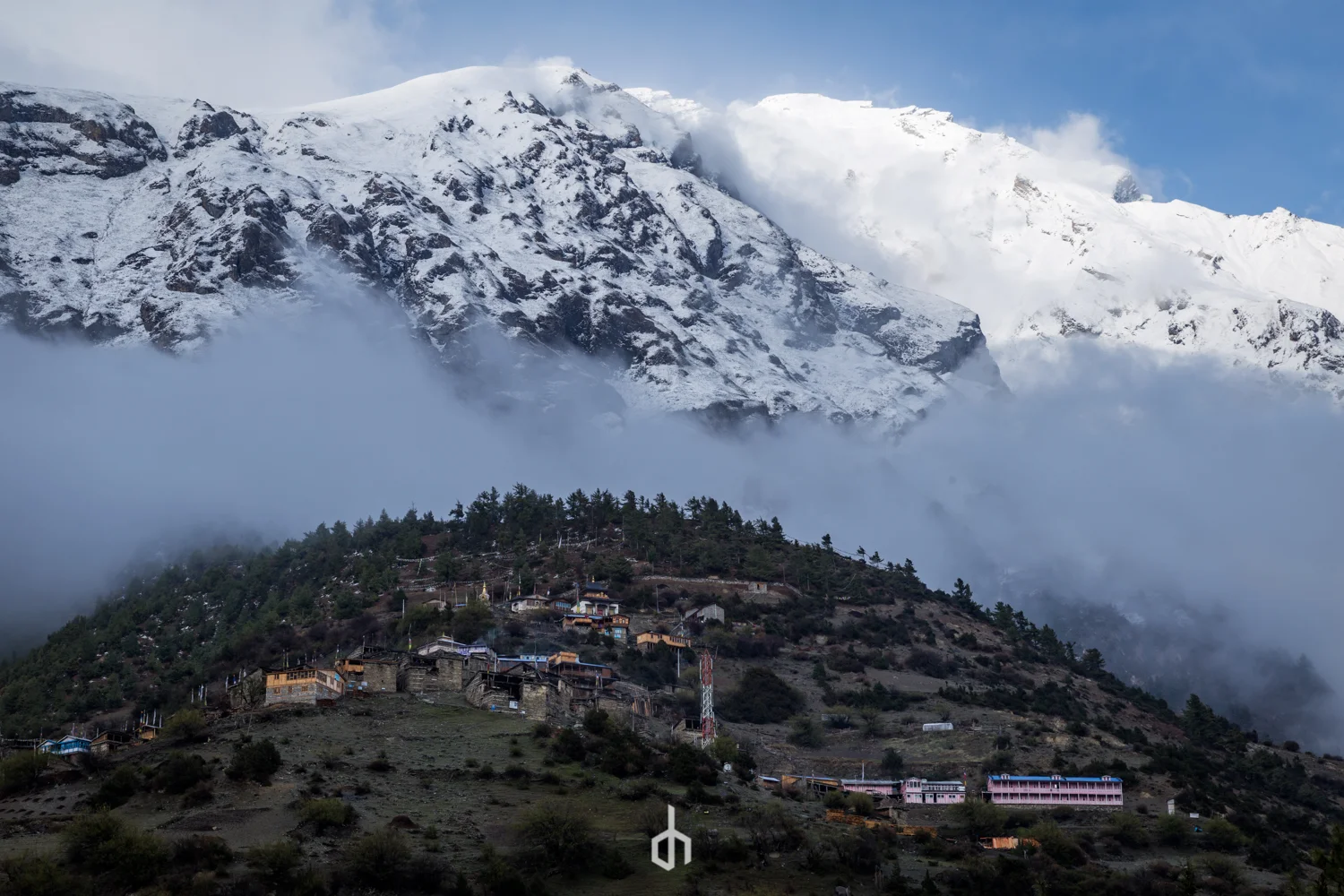 Snow-covered mountains with clouds and fog, a hillside village at the base with buildings and trees.