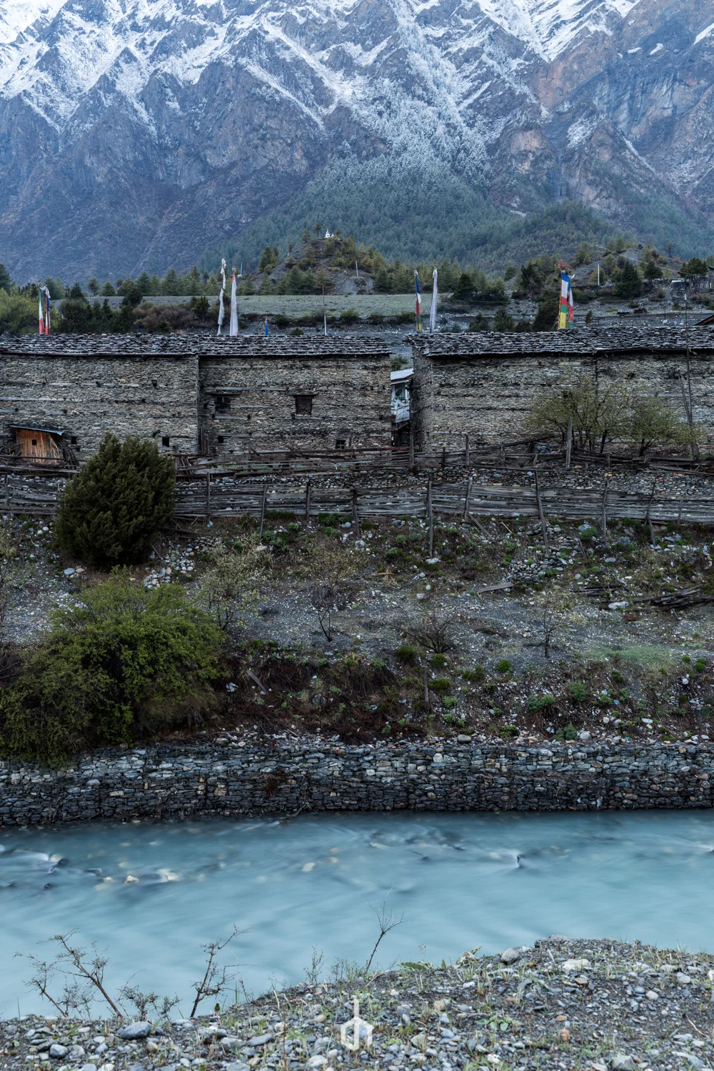 Traditional stone houses along a river with mountains in the background, some with snow, and colorful prayer flags on the hillside.