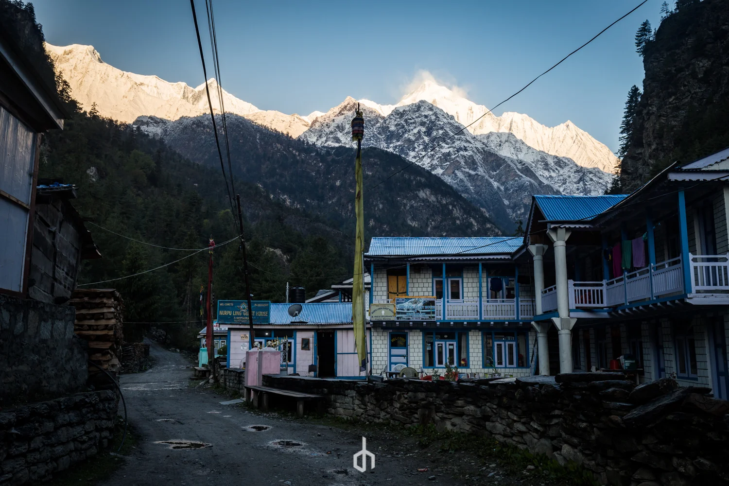 A mountain village with snow-capped peaks in the background, houses with blue roofs, and a dirt road in the foreground.