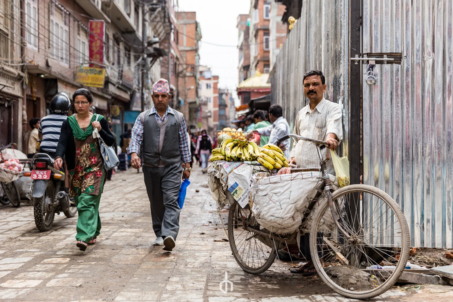 Street scene with a banana vendor selling bananas from a bicycle cart, pedestrians walking along the cobblestone street, and buildings lining the street.