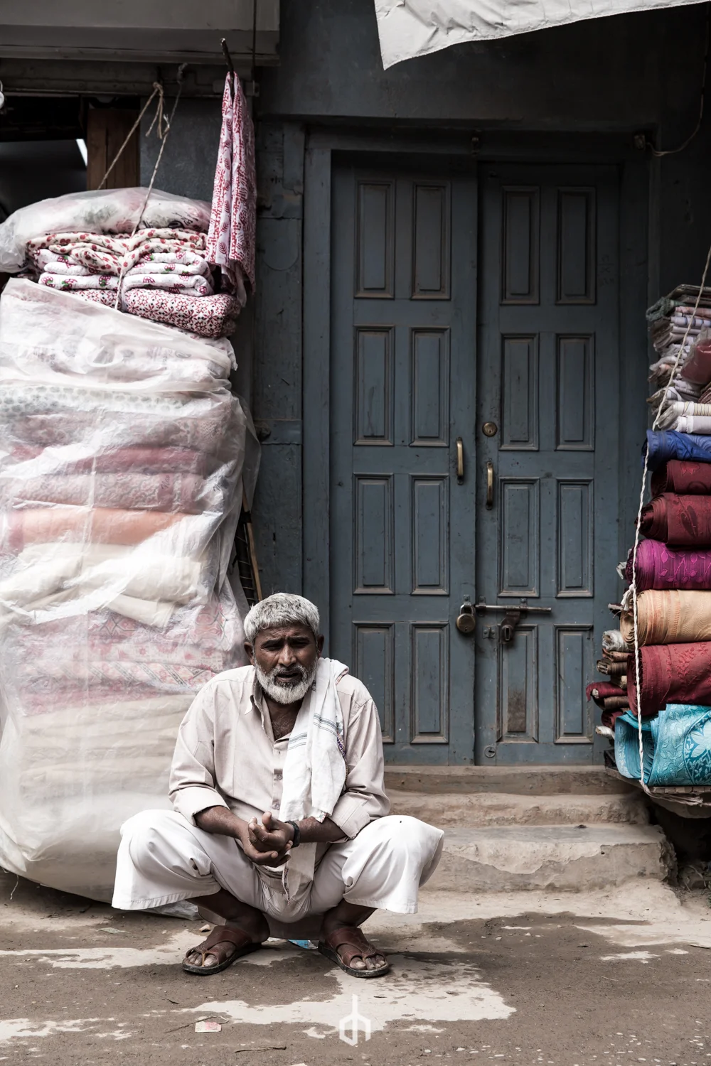 An elderly man with gray hair and beard, sitting cross-legged in front of a closed blue door. He is wearing beige traditional clothing with a white cloth draped over his shoulder and sandals. Behind him, there are stacks of fabric and blankets on eit