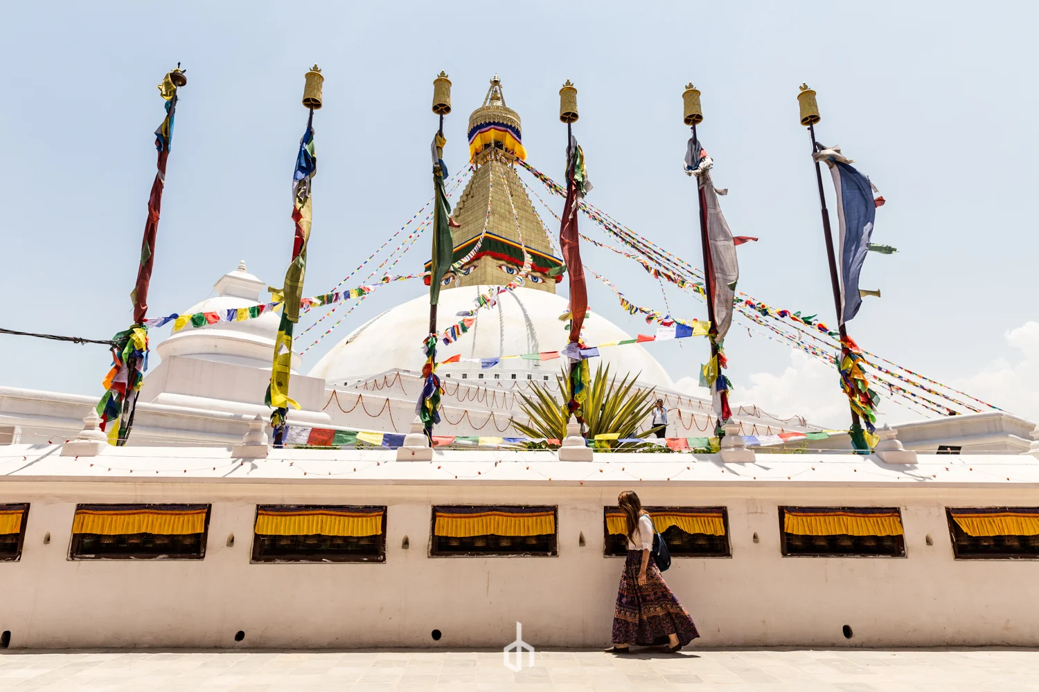 A woman walking past a white stupa with colorful prayer flags hanging around it, featuring a golden spire with eyes painted on the structure, indicating a Buddhist religious site in Nepal.