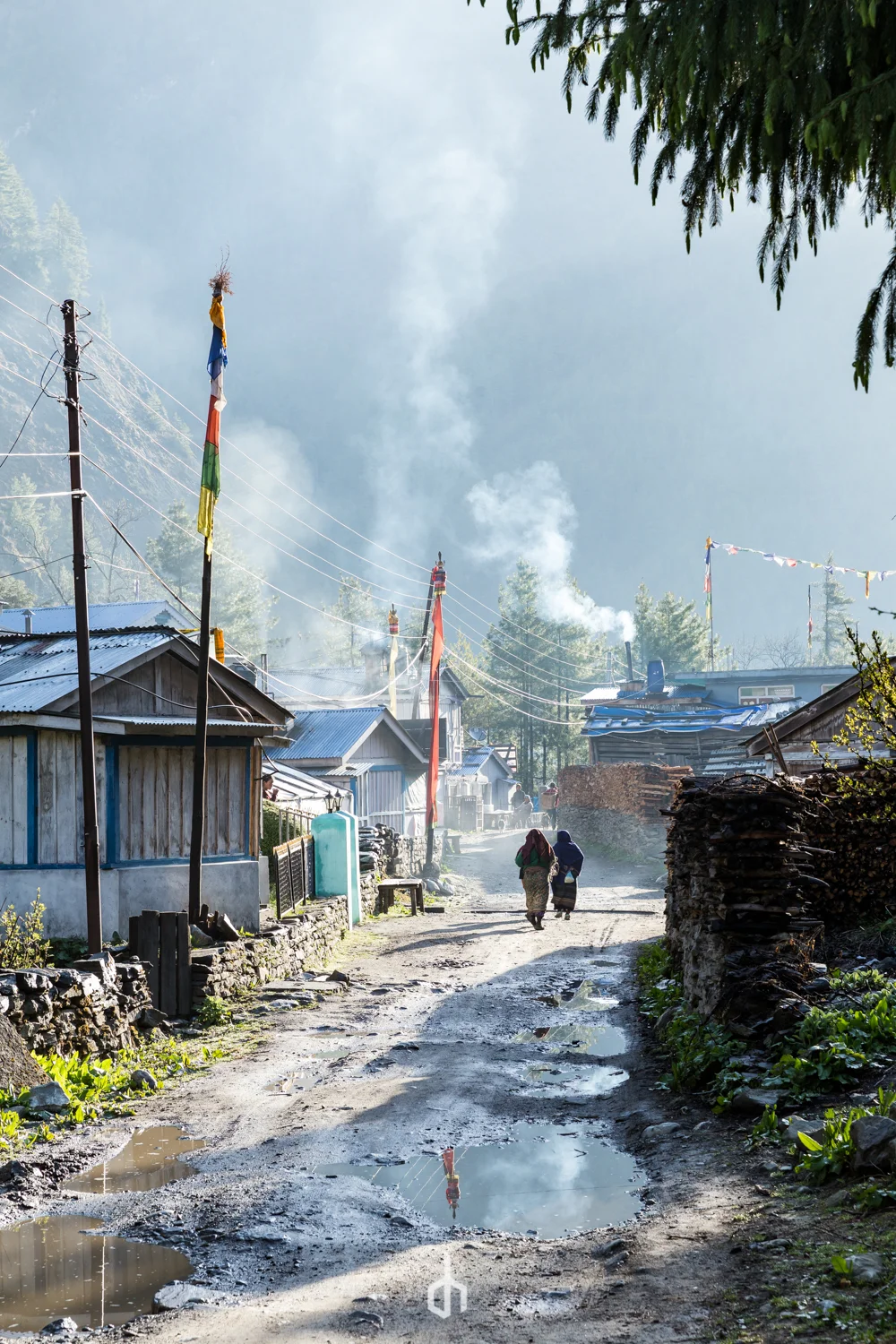 Dirt road in a rural village with puddles, lined with wood and metal houses, colorful prayer flags and smoke rising from chimneys, with two people walking away and trees in the background under a cloudy sky.