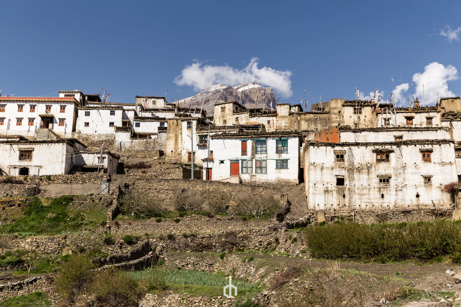 Hillside village with white and beige buildings, mountain in background, blue sky with clouds.