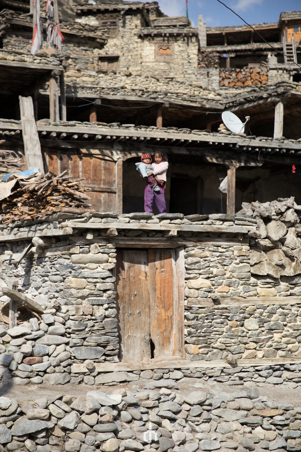 A woman and child standing on a stone platform in front of a rustic, stone and wood house in a mountainous area.