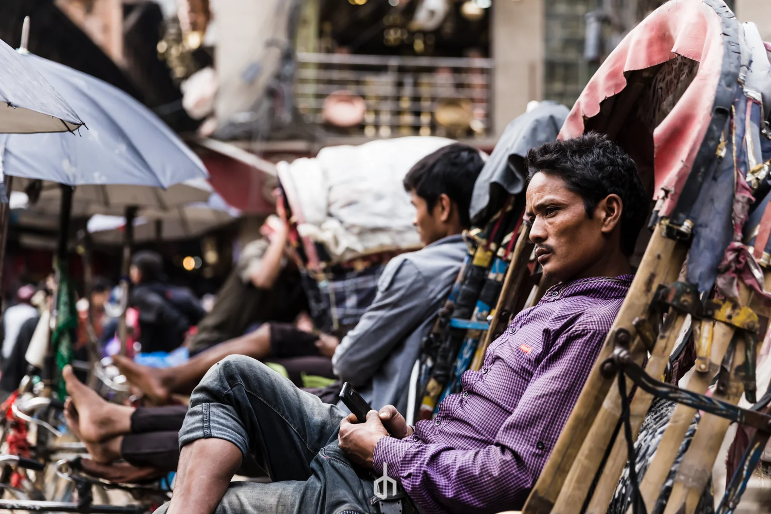 Man sitting on a bench at a busy street market, holding a smartphone, surrounded by pedestrians and market stalls with umbrellas.