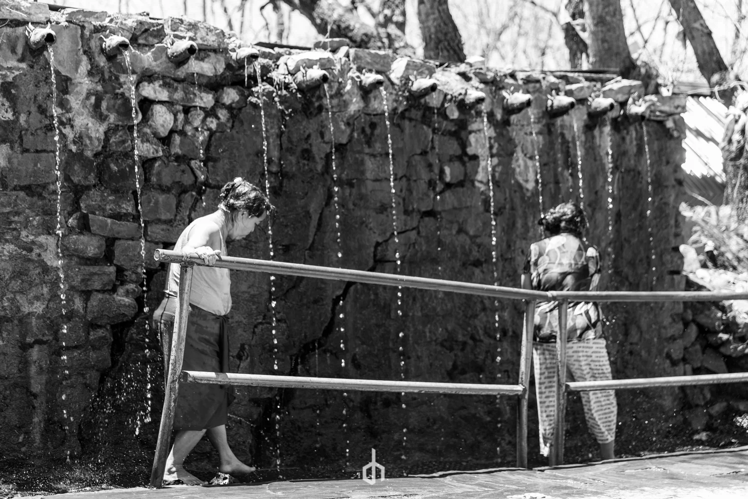 Two women stand under outdoor water fountains with multiple spouts flowing water. One woman is barefoot, washing her feet, while the other is adjusting her clothes or preparing to wash. The scene is outdoors with trees in the background.