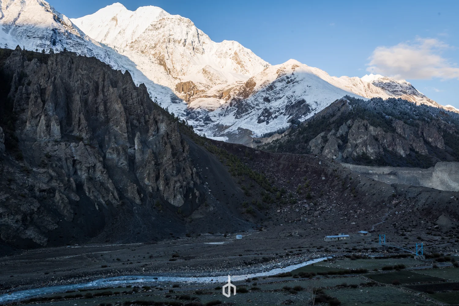 Snow-capped mountains with rocky cliffs and a valley with a river and small structures.