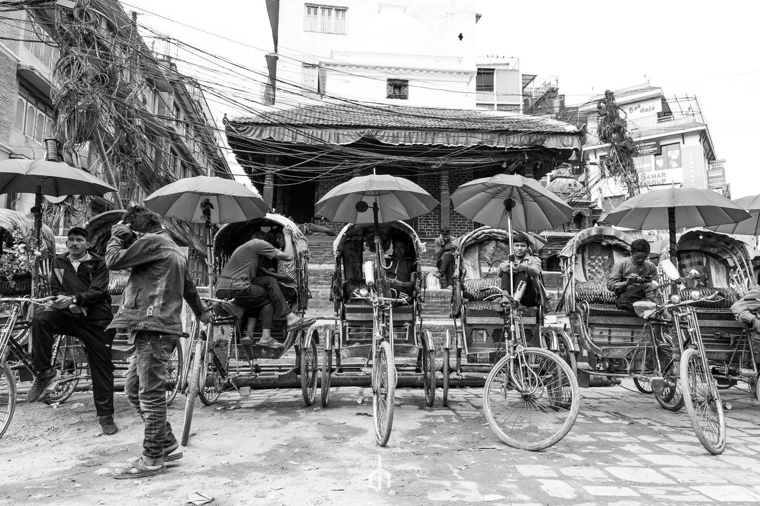 A black and white photo of a busy street scene with several rickshaws and people gathered, some sitting or standing under umbrellas, in front of a brick building with stairs leading up to it.