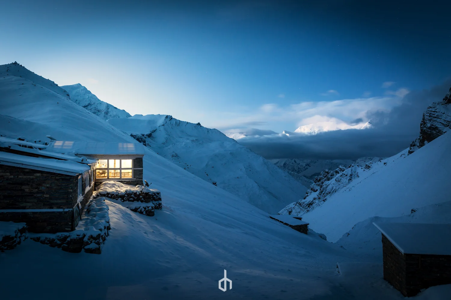 Snow-covered mountain landscape at dusk with two wooden cabins, one with illuminated windows, in a remote valley surrounded by steep peaks and cloudy sky.