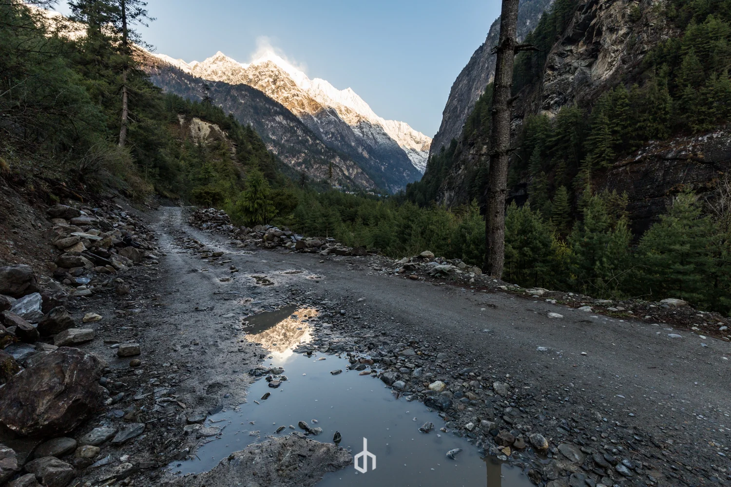 A rugged dirt mountain road with puddles reflective of the snow-capped peaks and pine forests surrounding the area.