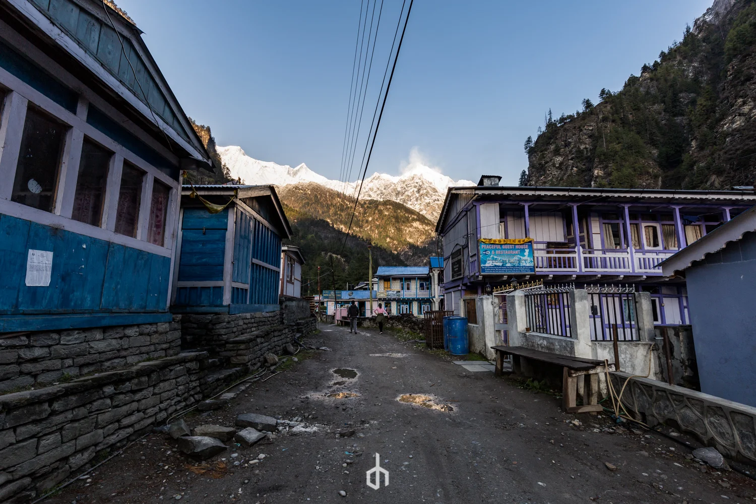 A dirt street in a mountain village with colorful wooden houses and snow-capped peaks in the background.