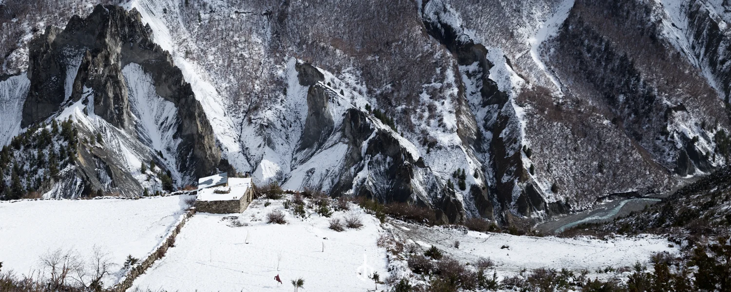 Snow-covered mountains with steep slopes and rugged cliffs, a small stone building with a snow-covered roof, and a winding river in the valley below.
