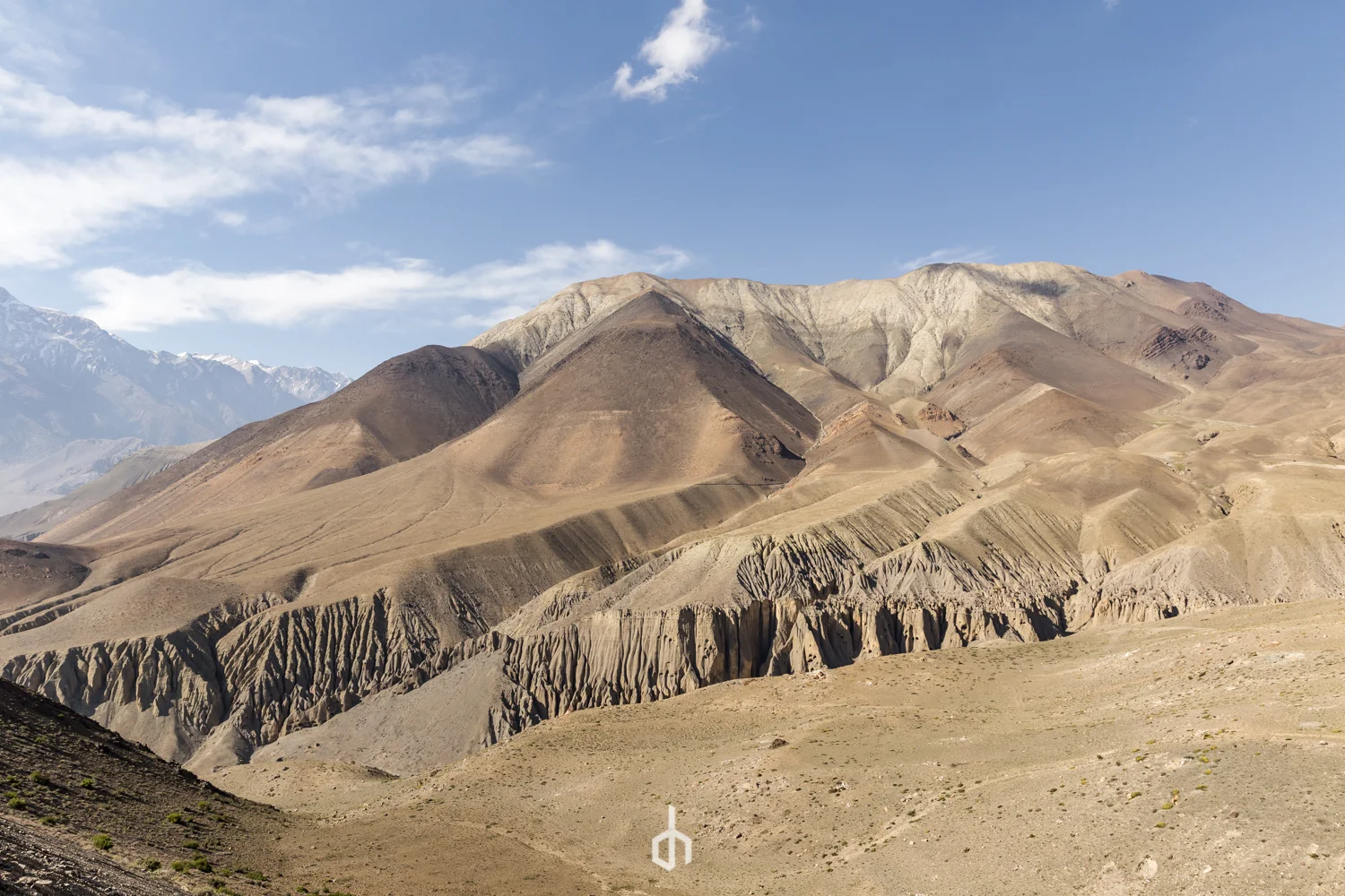 Mountain landscape with rugged, barren slopes, and distant snow-capped peaks under a blue sky with scattered clouds.