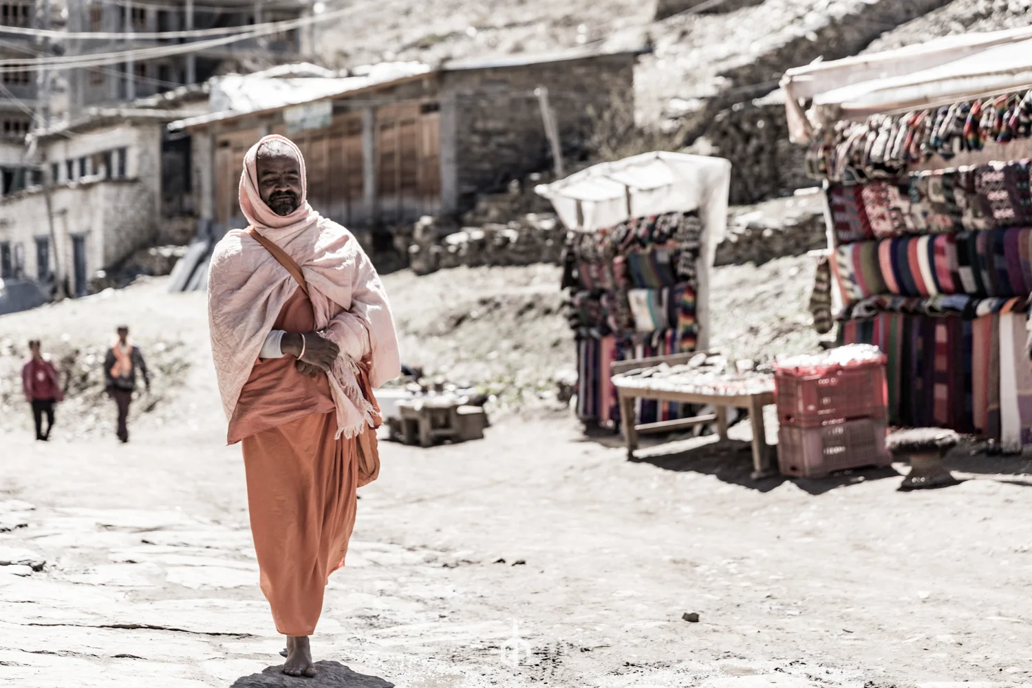 Man dressed in traditional saffron robes walking barefoot on a dirt road, with market stalls and other people in the background in a hilly, rural area.