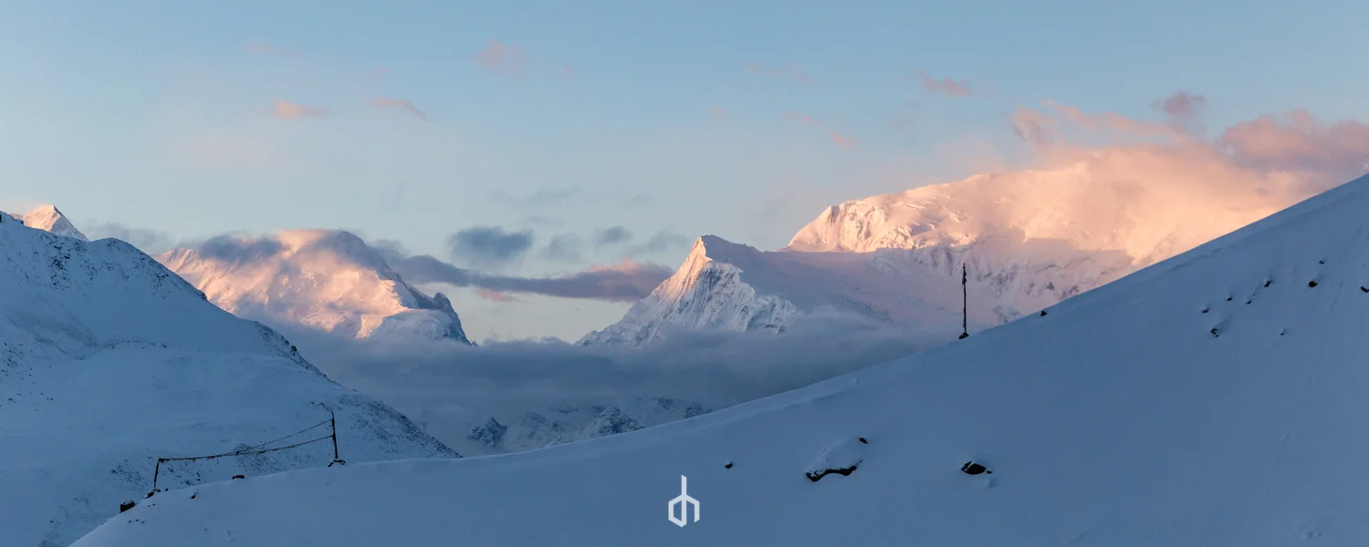 Snow-covered mountain range with clouds and pink hues from the setting or rising sun.