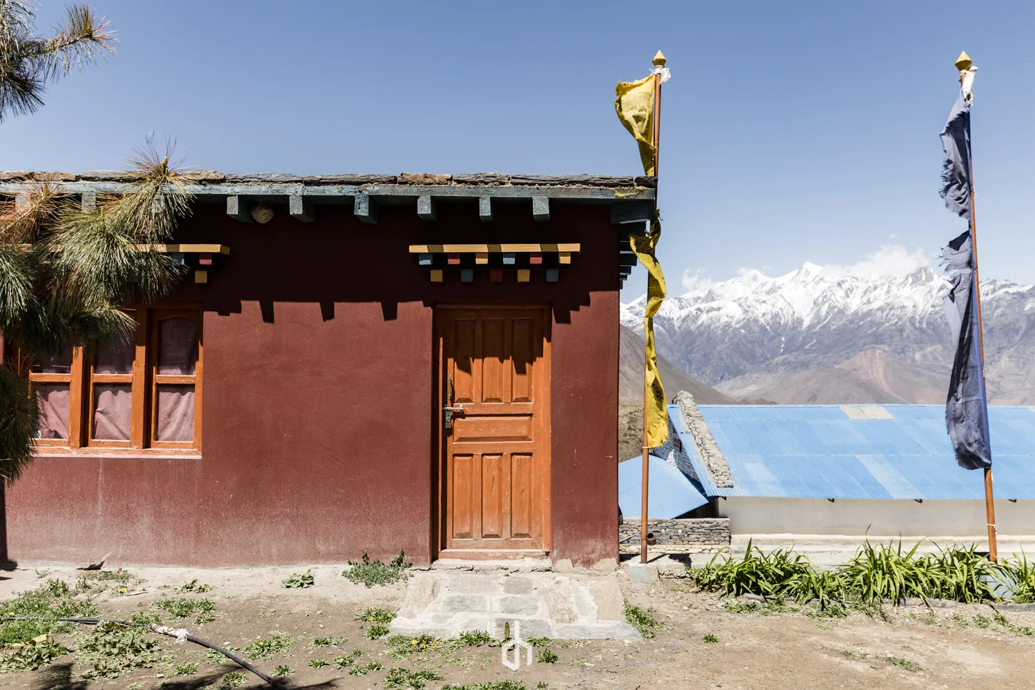 A small red building with a wooden door and window, flags on poles, and snow-capped mountains in the background.