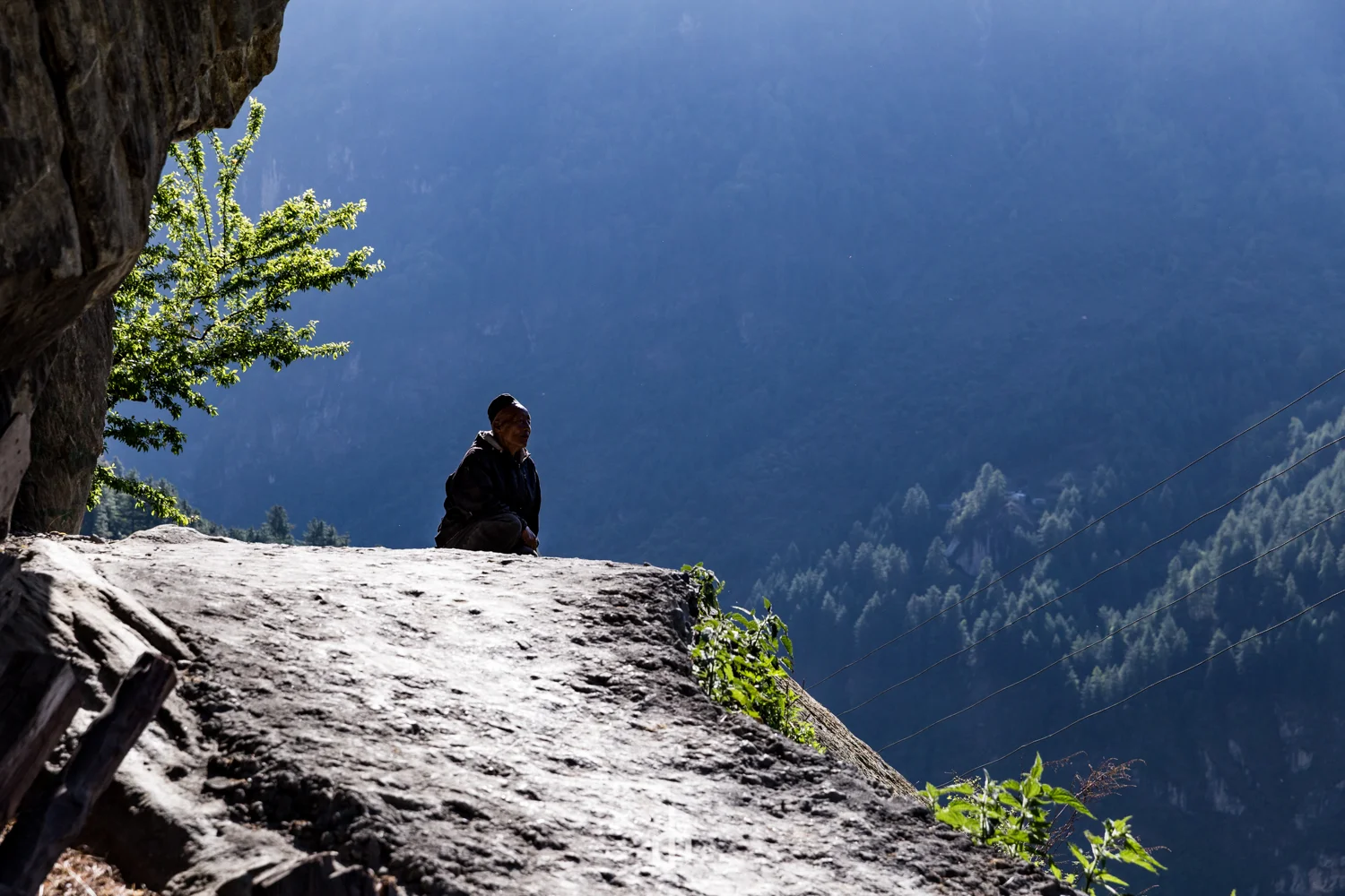 A person sitting on a large stone or cliff edge with a mountainous landscape in the background. The person appears to be looking into the distance, surrounded by greenery and forested mountains under a clear sky.