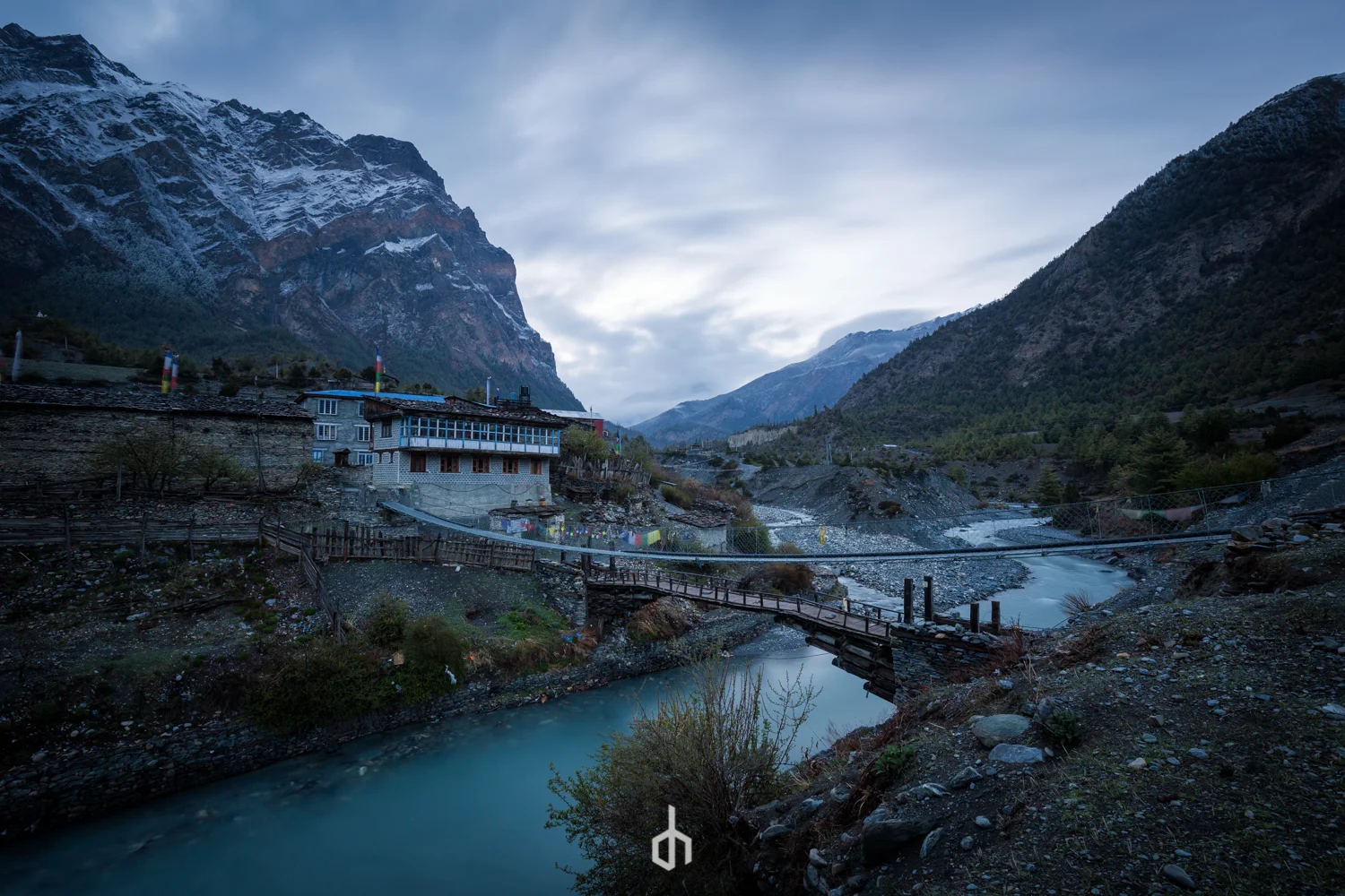 Scenic view of a mountain valley with a river, a bridge, and a small building on the hillside during early evening or dawn.