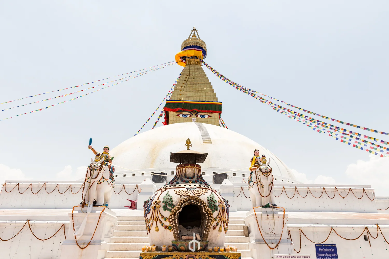 The image shows a large white stupa with colorful prayer flags hanging from its top. The stupa has a pair of painted eyes on it, symbolizing Buddha, and is decorated with intricate patterns and small statues. There are two mannequins on horseback at 