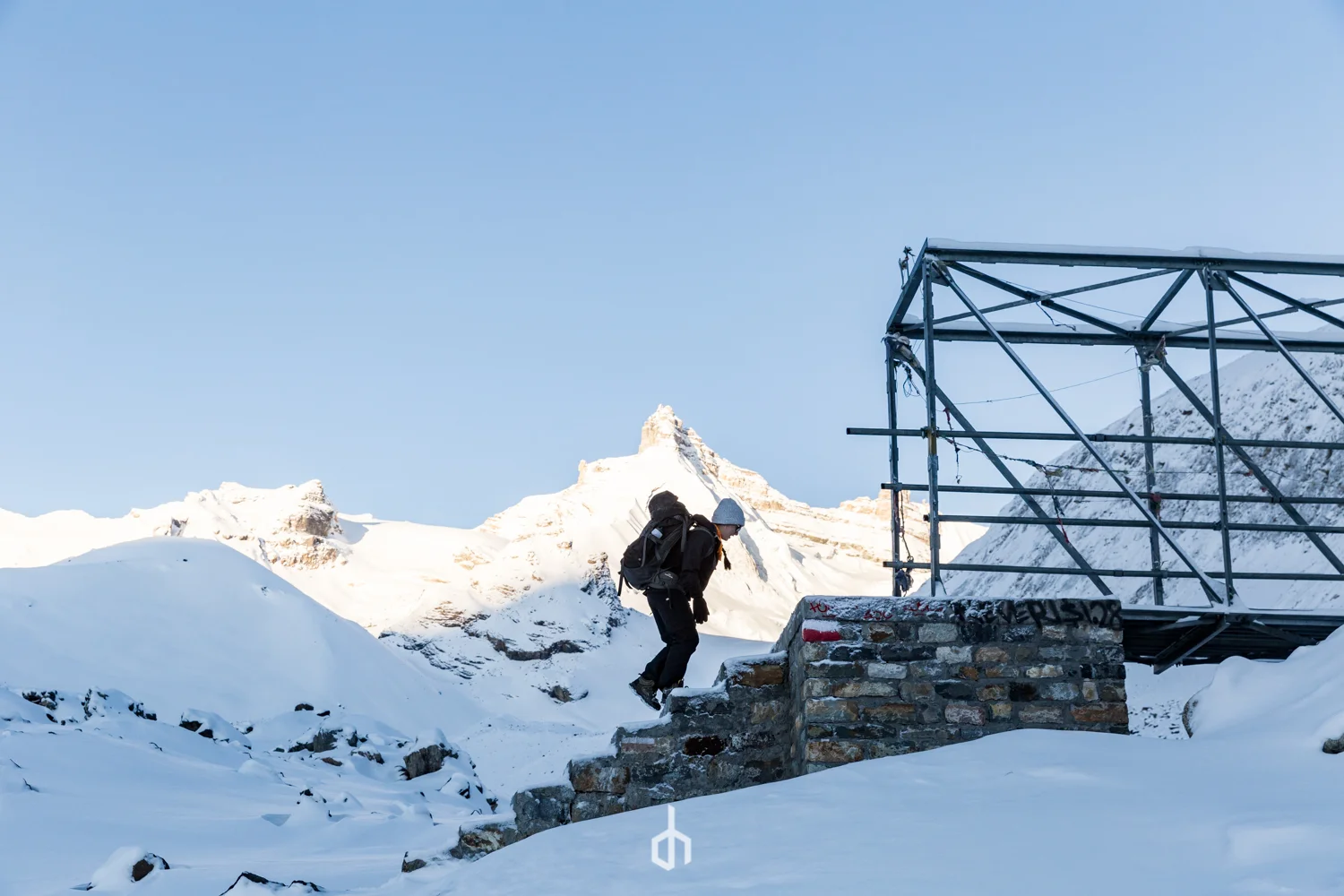 A person climbing up snow-covered steps in a mountainous area during winter with snow-covered peaks and scaffolding nearby.