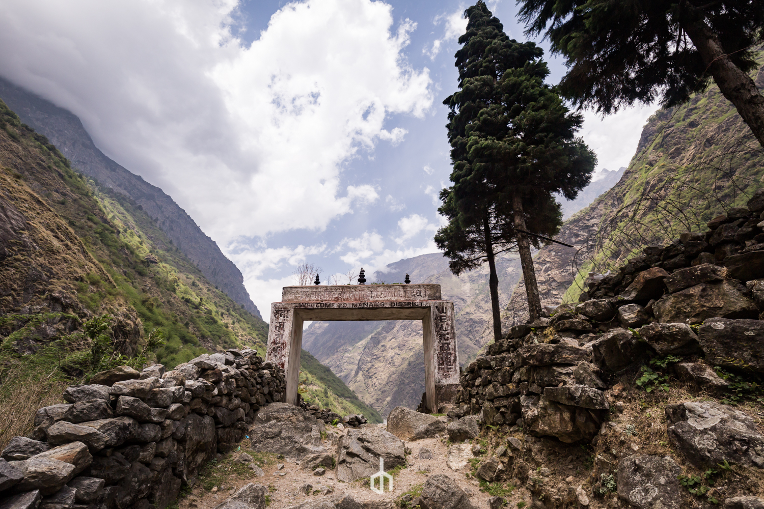 Mountain pass with rocky walls on either side, a stone archway in the middle with inscriptions, trees by the rocks, and a partly cloudy sky.