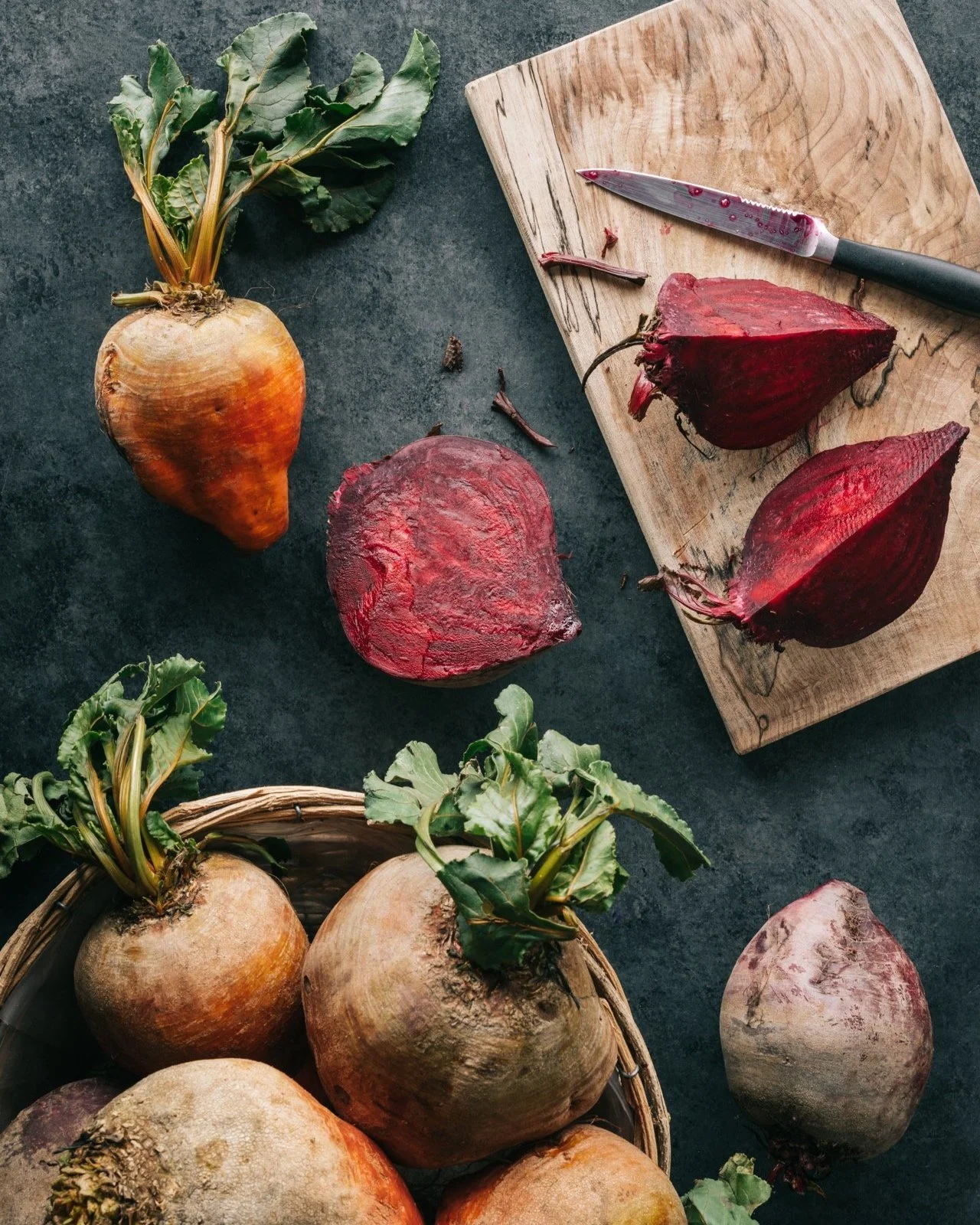Fresh beets and turnips with green leaves, some sliced on a wooden cutting board, on a dark kitchen countertop.