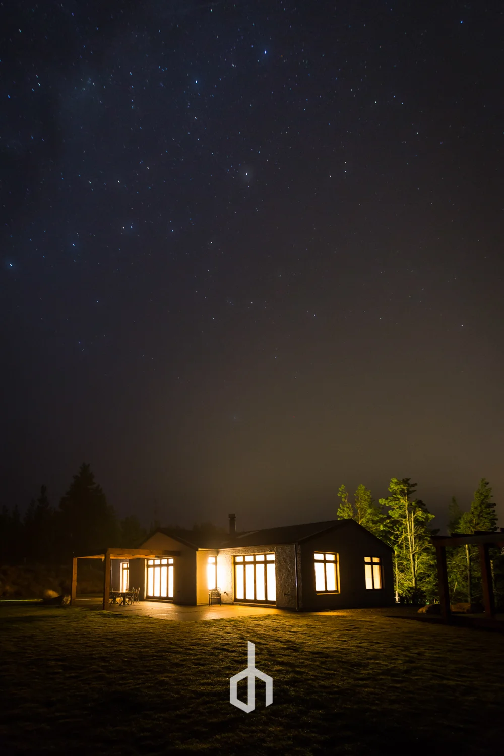 A house at night with lights on, surrounded by trees and a starry sky above.